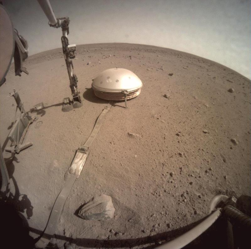 Marsquake: Fisheye lens view of white dome with data cord running to it, resting on reddish soil, and part of robotic arm.