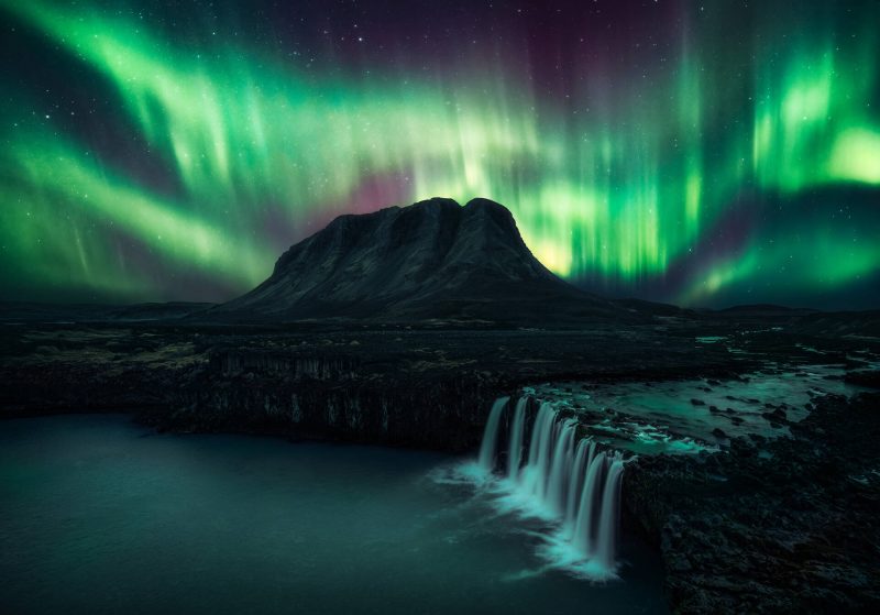 Distant mountain, foreground waterfall, and green and purple vertically striated bands of aurora in the sky.