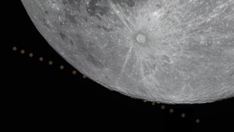 Moon with bright crater near limb and dot of orange circle repeating behind it.