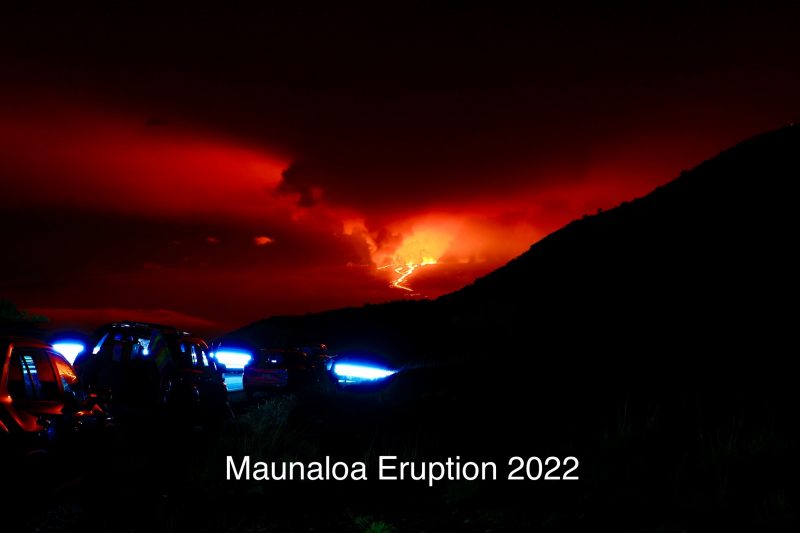 Cars in foreground lit by passing light, background of red clouds over brilliantly glowing lava flows.