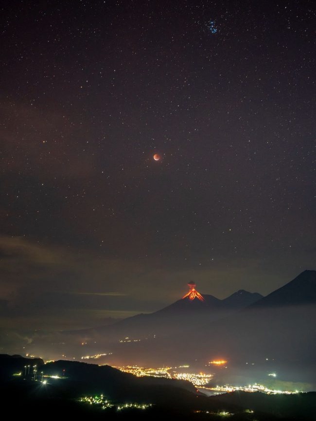 City lights in valley, with reddish volcano behind and reddish lunar eclipse high above.