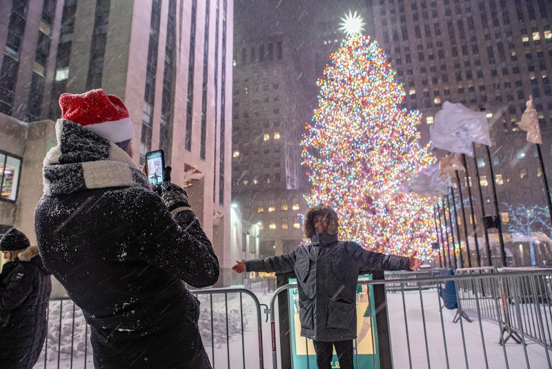 People taking photos in front of tall brilliantly lit conical tree, in a snowstorm.
