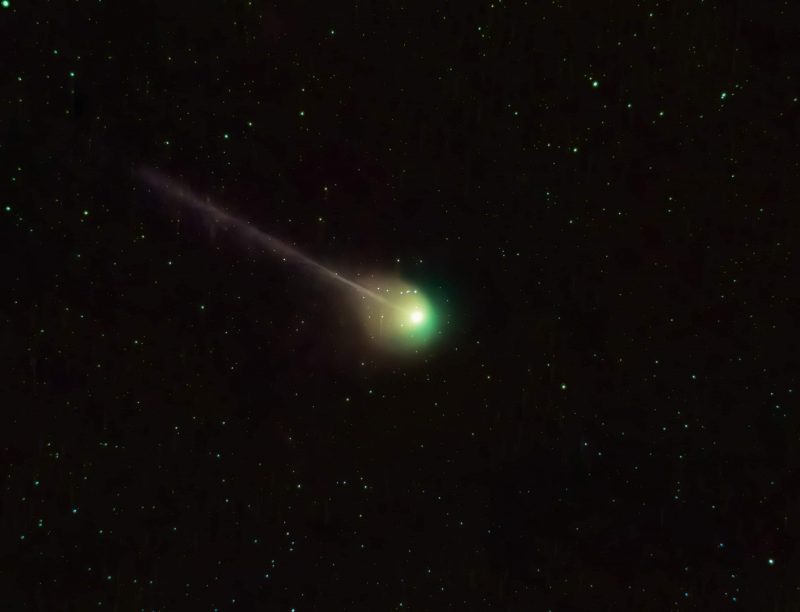 Bright fuzzy green object in center of star field with thin, straight streamer going to the upper left.
