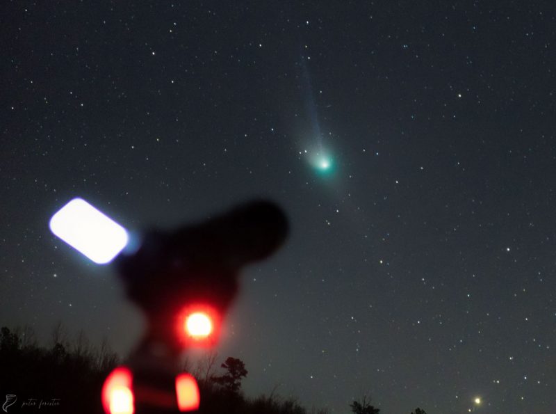 Telescope blurry in foreground with image of large green comet in sky behind.