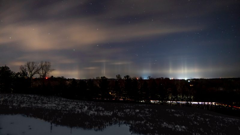 Cloudy night with vertical pillars of light coming up from distant town.