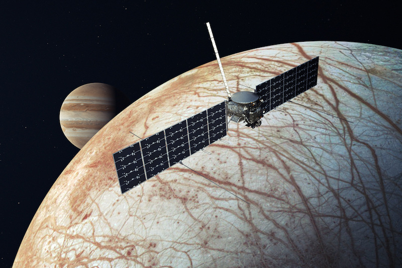 Europa Clipper: Spacecraft with 2 large solar panels near an off-white moon with many brown cracks on its surface.