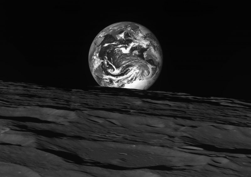 Black and white photo of full Earth beyond dim, gray cratered surface in the foreground.