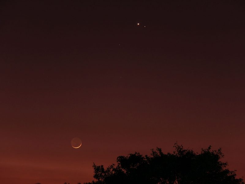 Dark reddish sky with thin crescent moon below and two small bright dots high above.