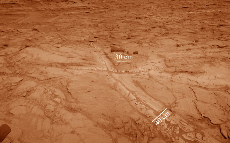 2 criss-crossing fractures in reddish rock with lighter-colored edges. Marks indicating size.