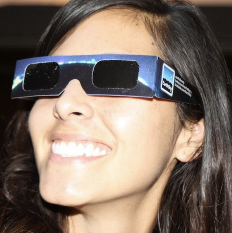 Closeup of smiling young woman's brightly sunlit face. She has on cardboard glasses with black lenses.