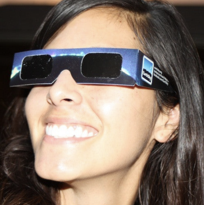 Closeup of smiling young woman's brightly sunlit face. She has on cardboard eclipse glasses with black lenses.