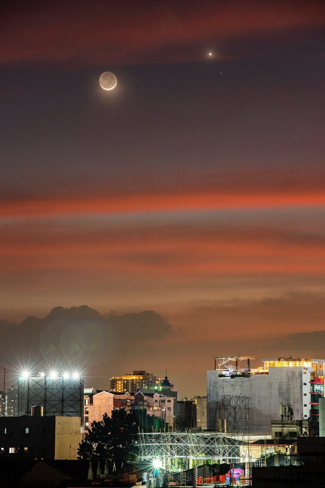 Venus, Saturn and crescent moon with earthshine above orange sunset clouds over a city.