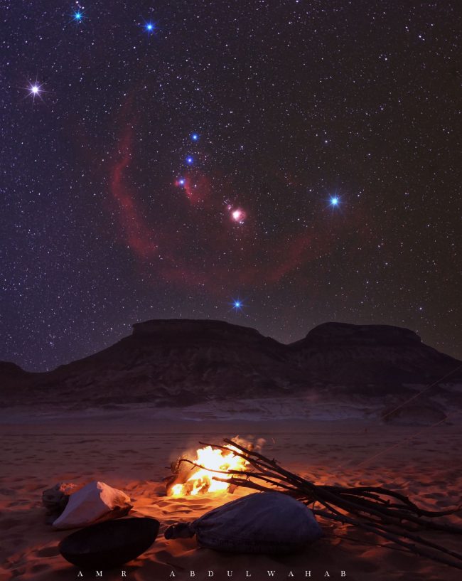 Orion, with a fuzzy red semicircular nebula looping through it, over a campfire.