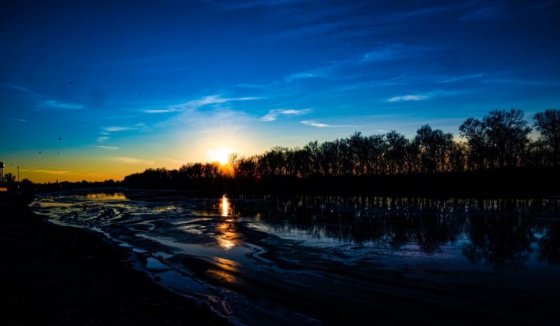 Brightest objects: Glow of setting sun behind trees under vivid deep blue sky, with icy river in foreground.