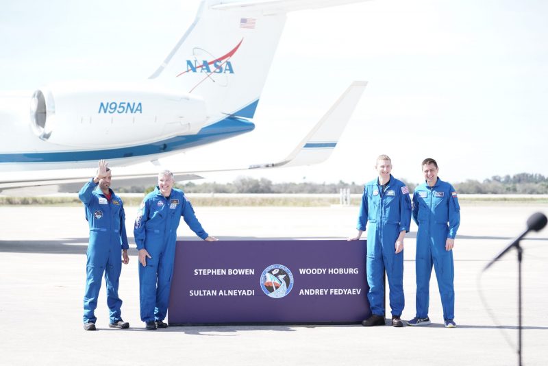 Crew-6: Four men in blue flight suits stand by a sign with their names with a NASA plane in the background.