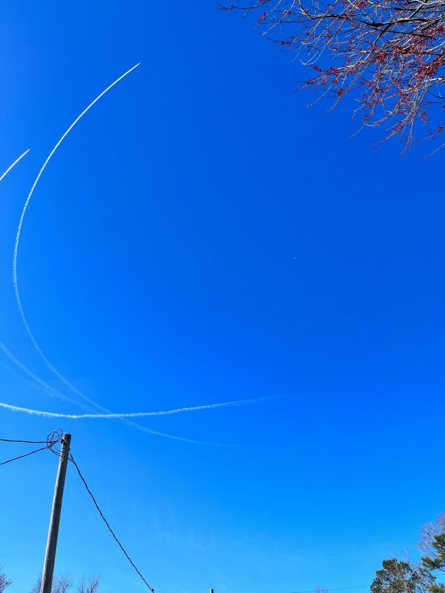 A wide-open view of blue sky with 3 arced contrails circling a small balloon.