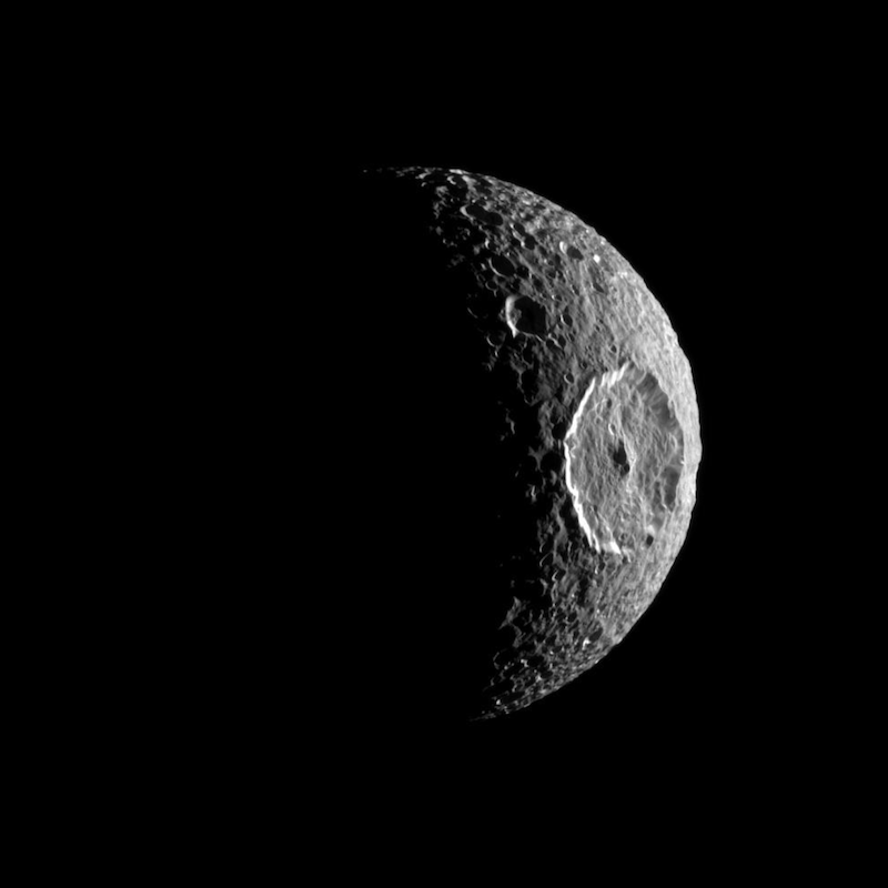 Saturn's moon Mimas: Heavily cratered moon-like object mostly in shadow, with 1 very large crater in sunlight.
