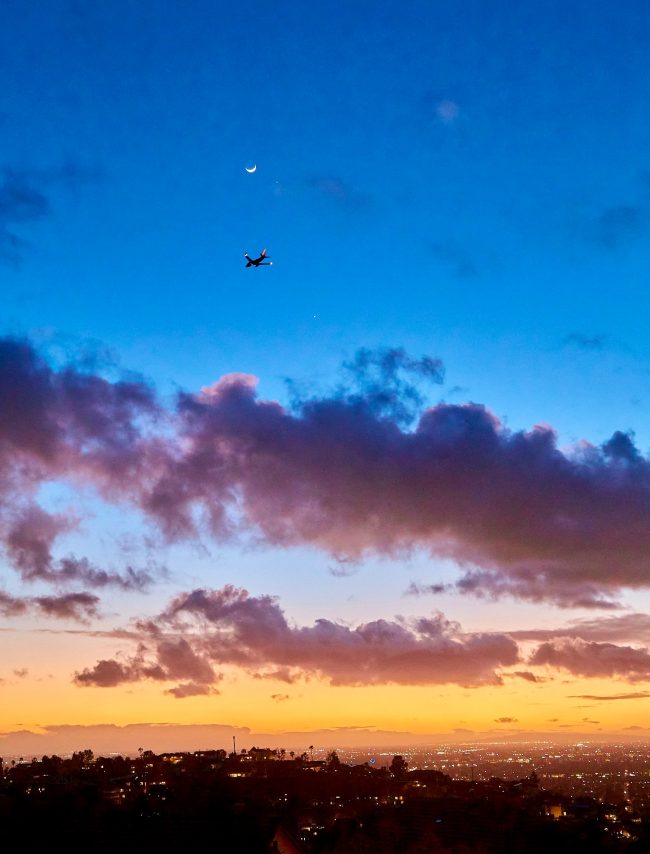 Sunset over cityscape with clouds and blue sky showing crescent moon, Venus and Jupiter and an airplane.