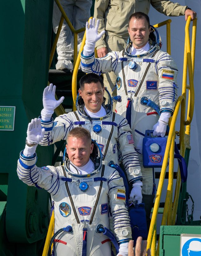 Stranded ISS crew: Space travelers going up a staircase to their rocket, and waving.