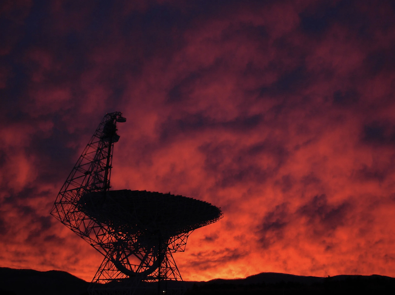 AI search for aliens: Radio telescope with large dish silhouetted against gloriously orange-red dark sunset sky.