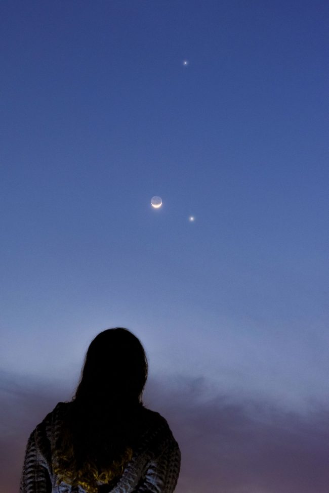 Person looking at a small crescent moon with two bright points nearby in medium-blue sky.