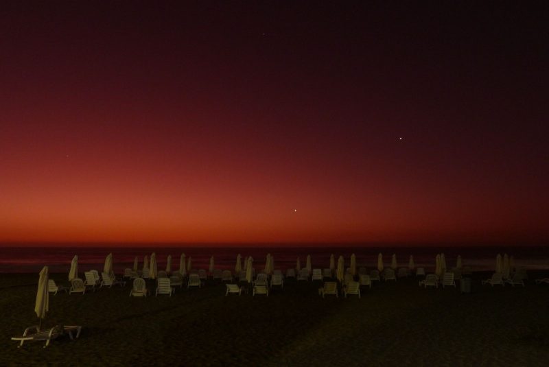 Empty beach chairs on a dark beach with an orange glow on the horizon and 2 dots in the sky.