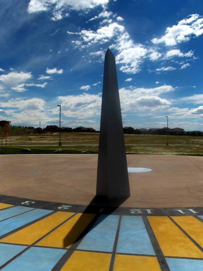 Tall black obelisk with shadow cast on radial blue and yellow checkerboard with times on it.