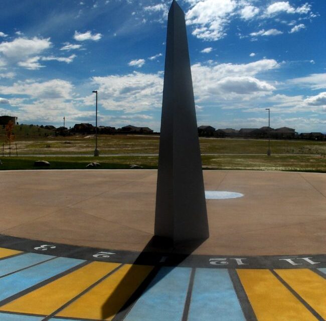 Tall, black obelisk with shadow cast on radial blue and yellow checkerboard with times on it.