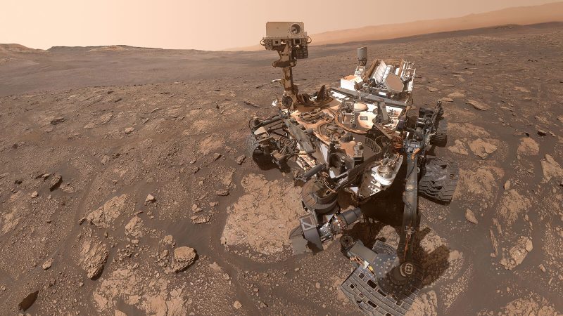 Complicated wheeled machine with a camera on top and a reddish desert as the background.