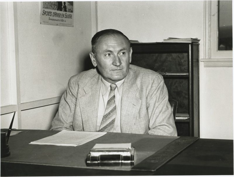 Man sitting behind a desk in suit and tie, with papers in front of him.