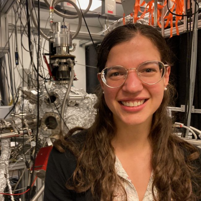 Young woman with long curly hair and glasses smiling with wires behind her.