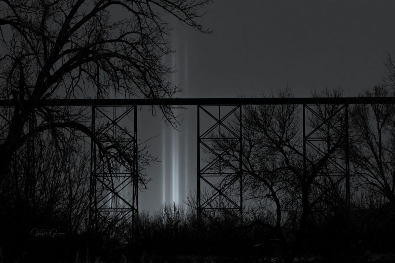 Light pillars in the background with a bridge and trees in the foreground.