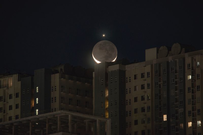 Tall building with crescent moon setting behind and a dot perched on top of the moon.
