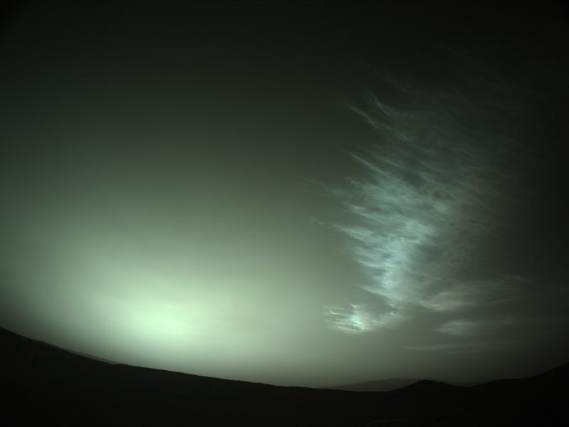 Noctilucent clouds on Mars: In a dark sky, a greenish light on the horizon and tendrils of high, light-colored clouds on the right.