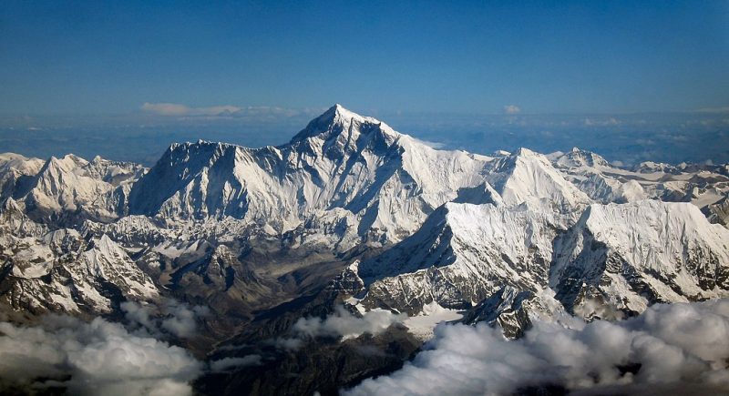 Mount Everest: Aerial view of range of sharp-pointed snow-covered mountains beneath blue sky with clouds far below the peaks.