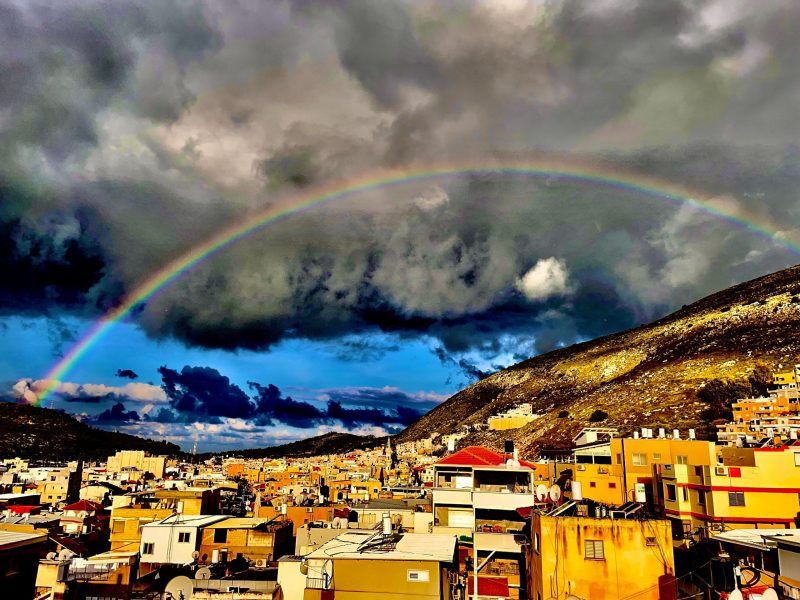 Rainbow arcing over a city brightly lit by the sun with dark clouds behind.