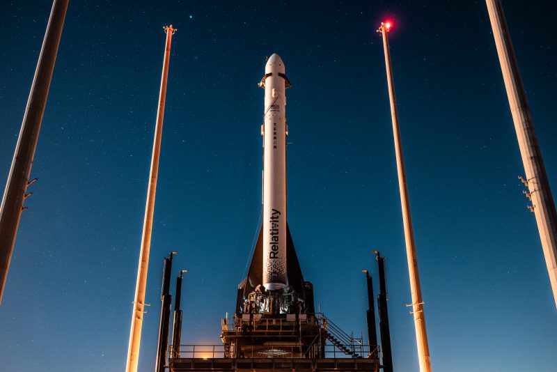 3D-printed rocket: White rocket, dark blue sky, tall thin poles with red lights on top of them.