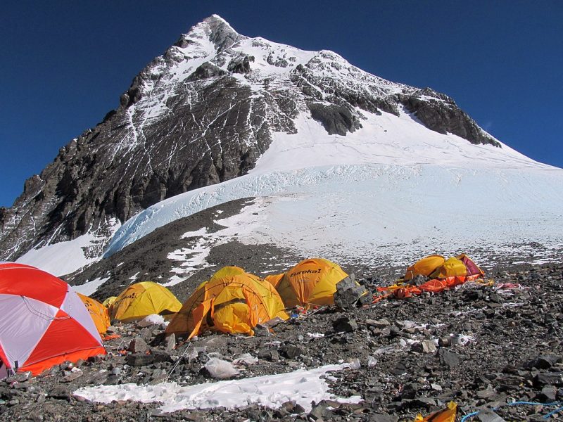 Rocky, snowy, steep mountain peak in background with yellow tents on rocky ground in foreground.