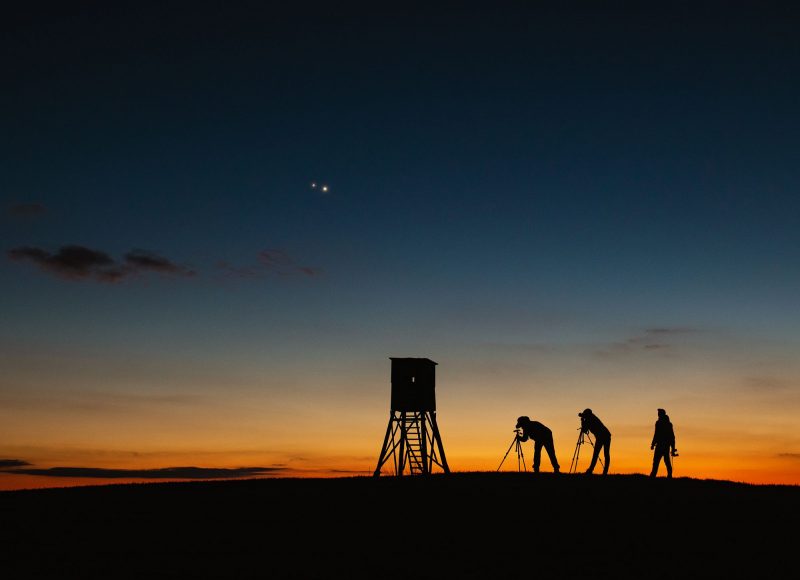 Three people with cameras on tripods, silhouetted in sunset and two close points of light above.