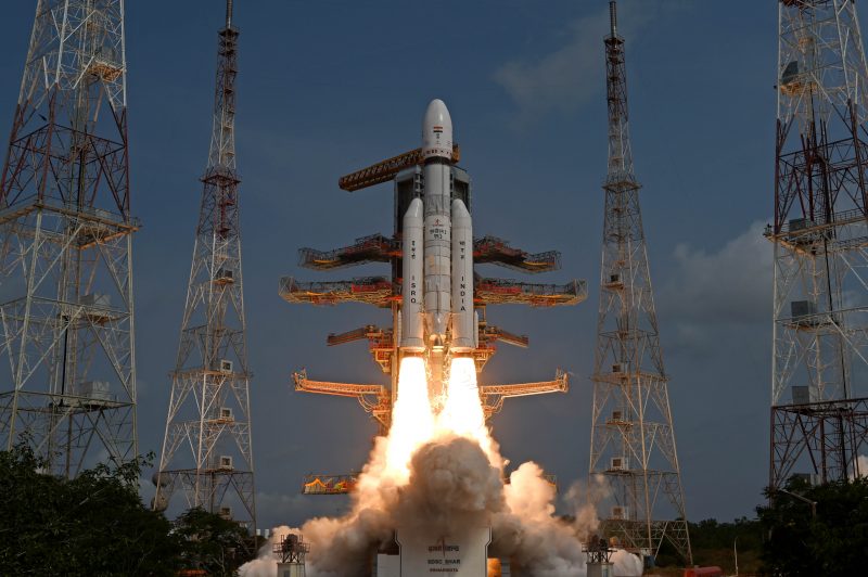 Indian rocket launch: The rocket is composed by 3 white cylindrical metal towers, there is a blue sky and fire smoke under the rocket.
