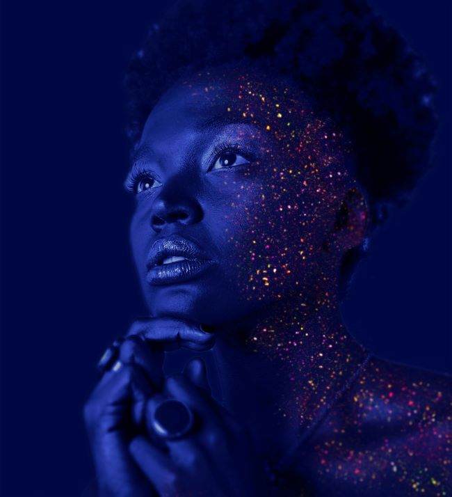 Black woman in a dark room with lit dots on one side of her face looking contemplative.