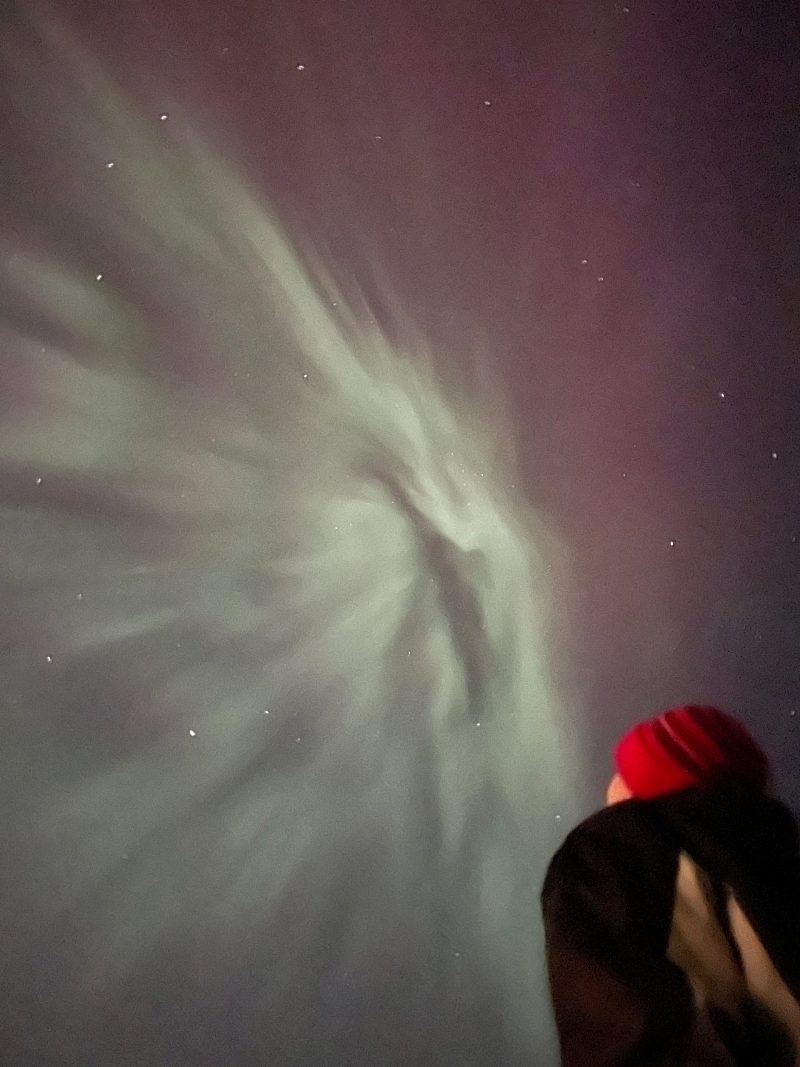 Man in coat and red knit cap looks up at pale green rays of aurora light in the sky.