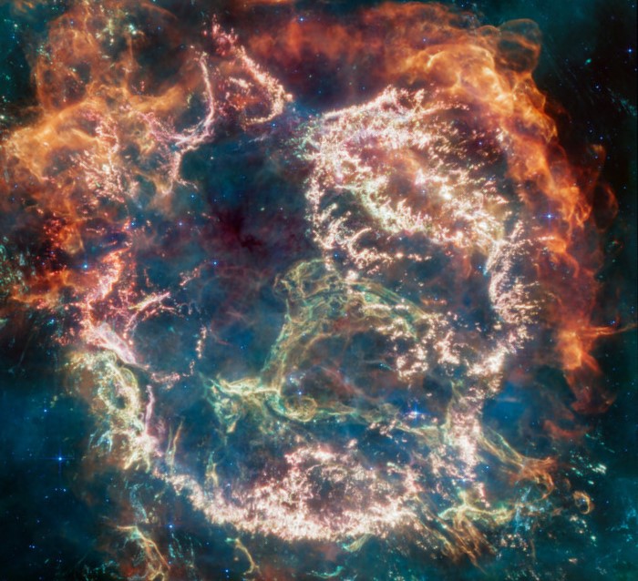 Supernova remnant: Roughly circular cloud of wispy strands colored from shades of orange to greenish blue.