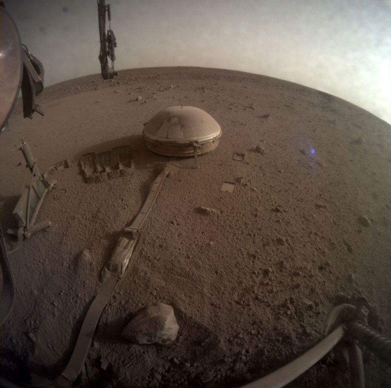 Metallic dome-like object with long ribbon attached to it, on reddish sandy and rocky terrain.