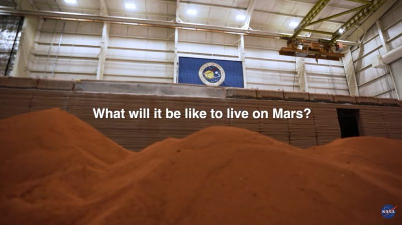 Mars habitat: Red piles of sand inside a large warehouse-type building with NASA flag high up on the wall.