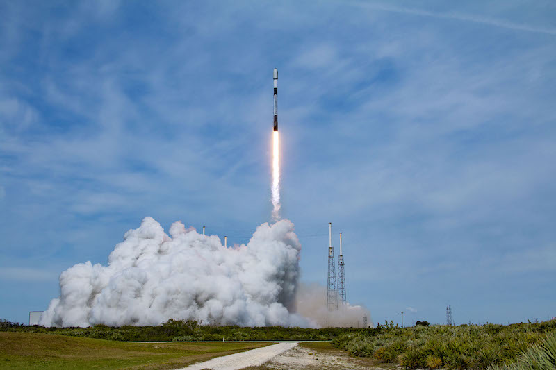 A white and black cylindrical vehicle launches upward into a blue sky, leaving behind clouds of smoke on the green ground.