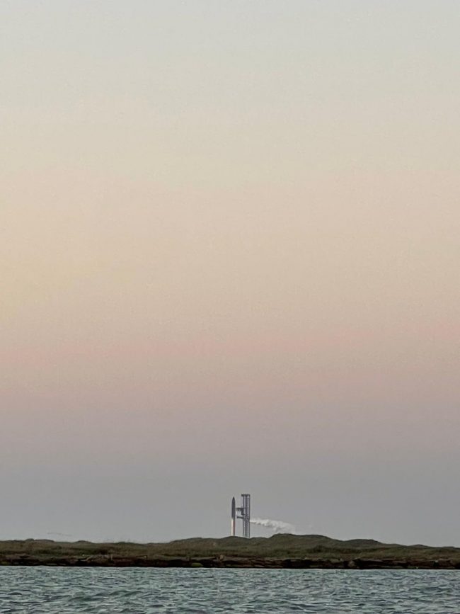 Pinkish blue sunrise colors and the vertical Starship rocket seen at a distance across a body of water.