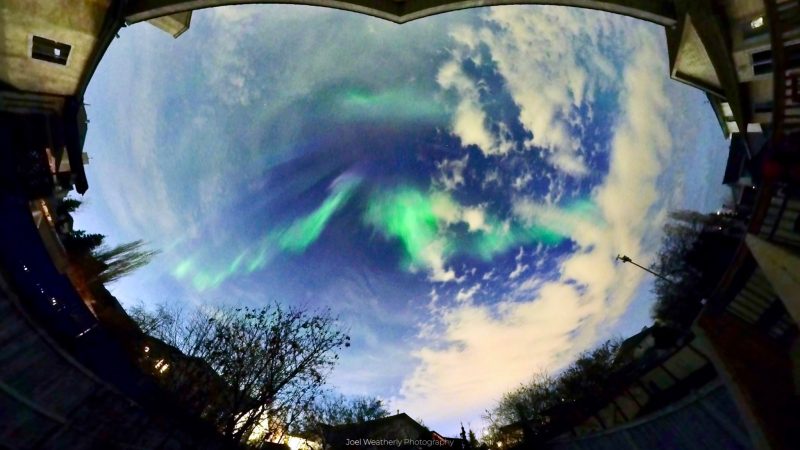 Panorama view of a bluish full sky with some clouds and green auroras.