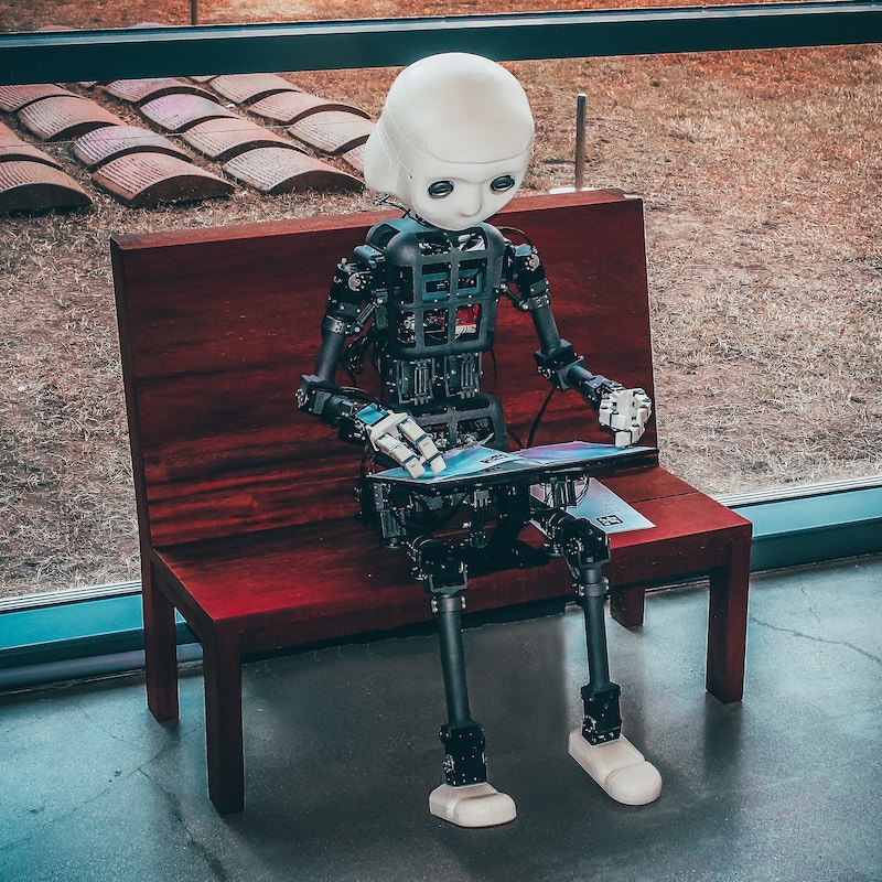 Artificial intelligence: Robot made of circuits with a white head and white shoes sitting on a bench reading a magazine.