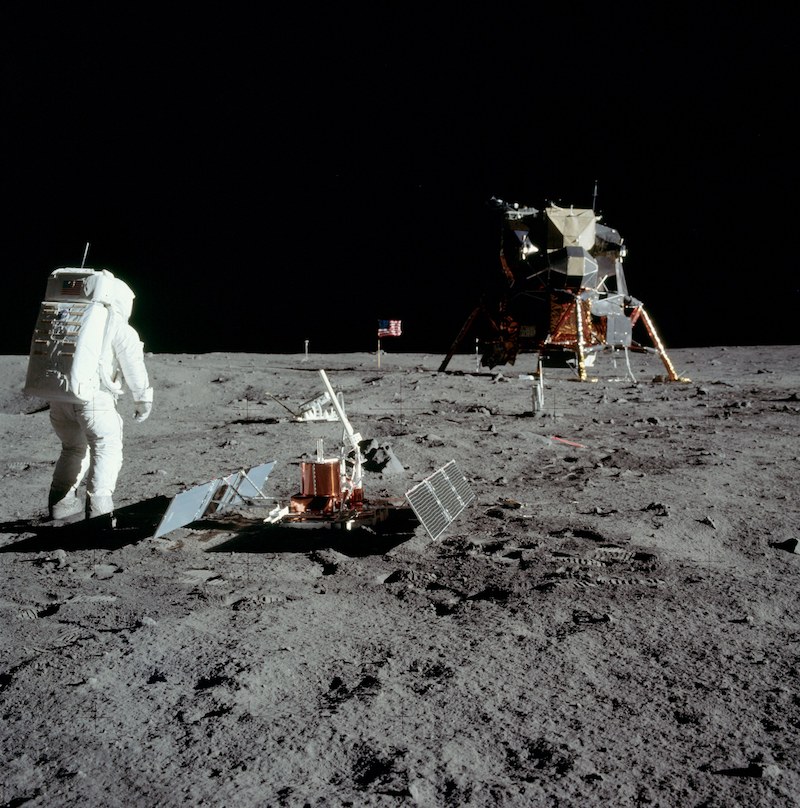 Astronaut in white space suit on gray terrain with landed spacecraft in distance.